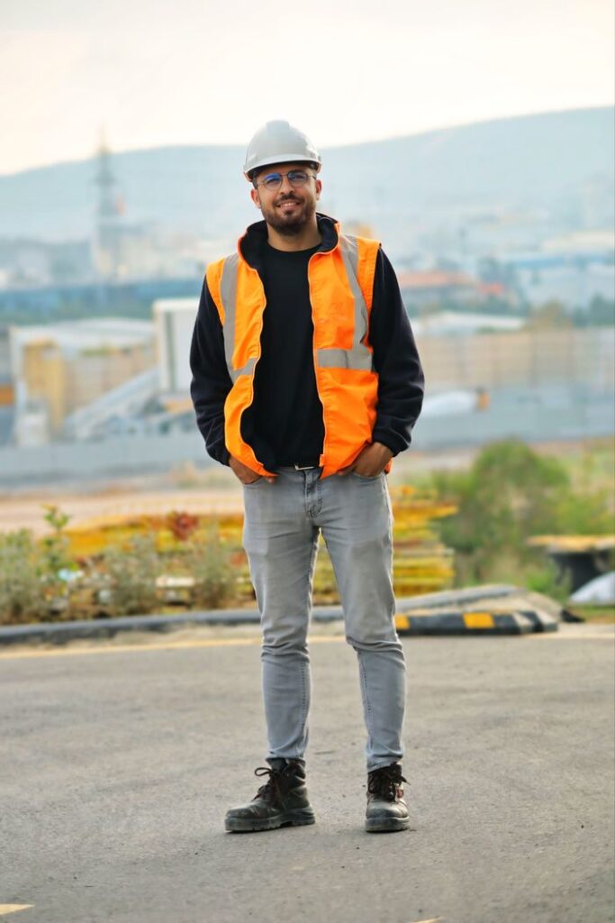 A construction worker stands confidently outdoors wearing safety gear and helmet.