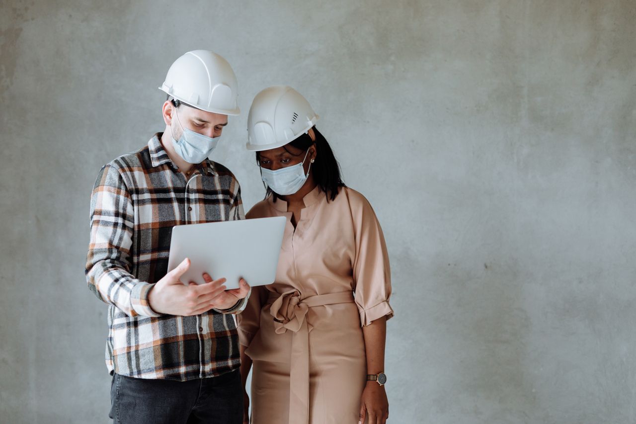 Two engineers wearing hard hats and masks review plans on a laptop.